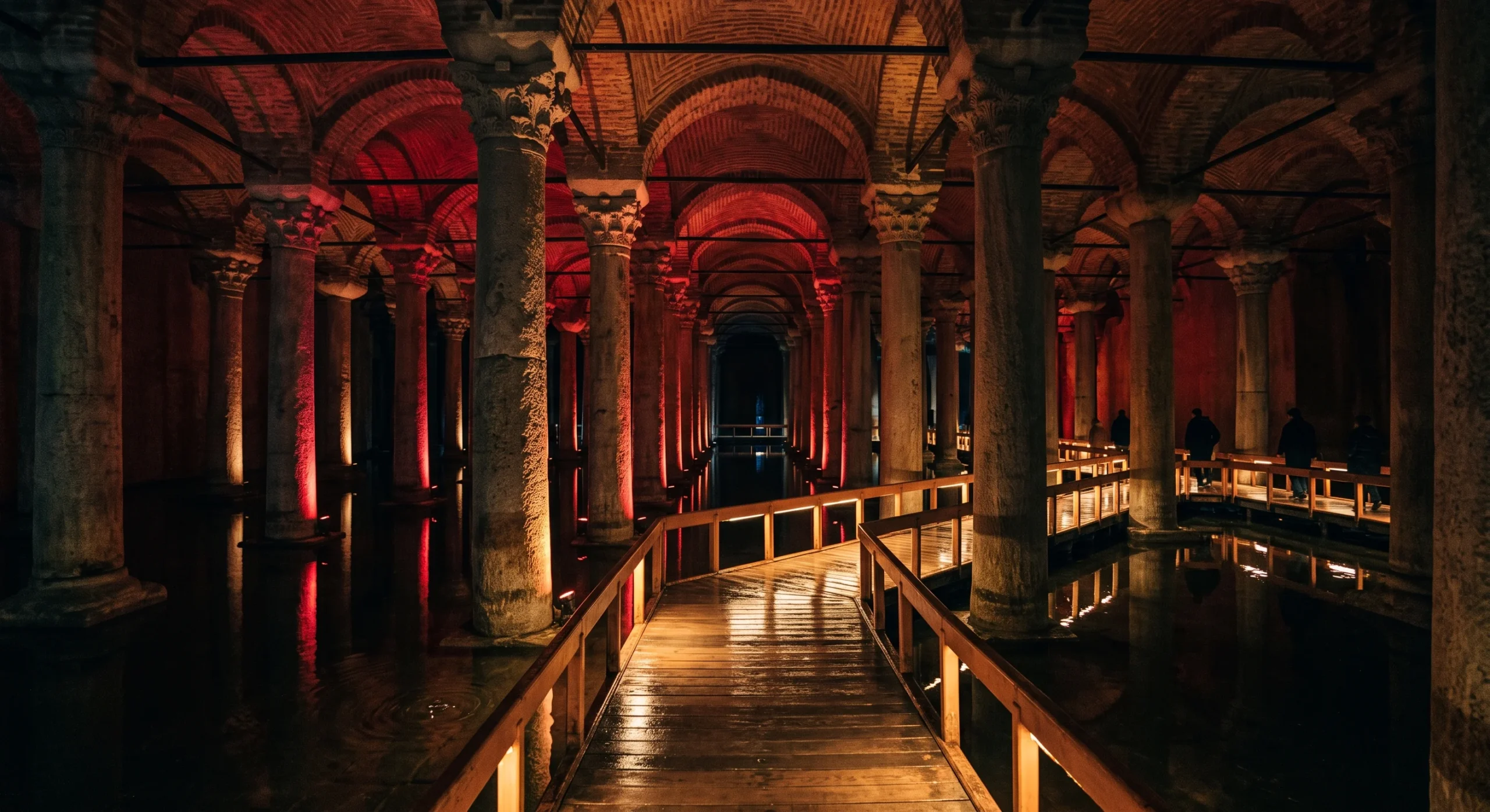 Atmospheric underground shot of the Basilica Cistern, rows of ancient stone columns rising from still dark water, dramatic uplighting in deep red and amber tones, a wooden walkway leading into the distance, moody subterranean composition.