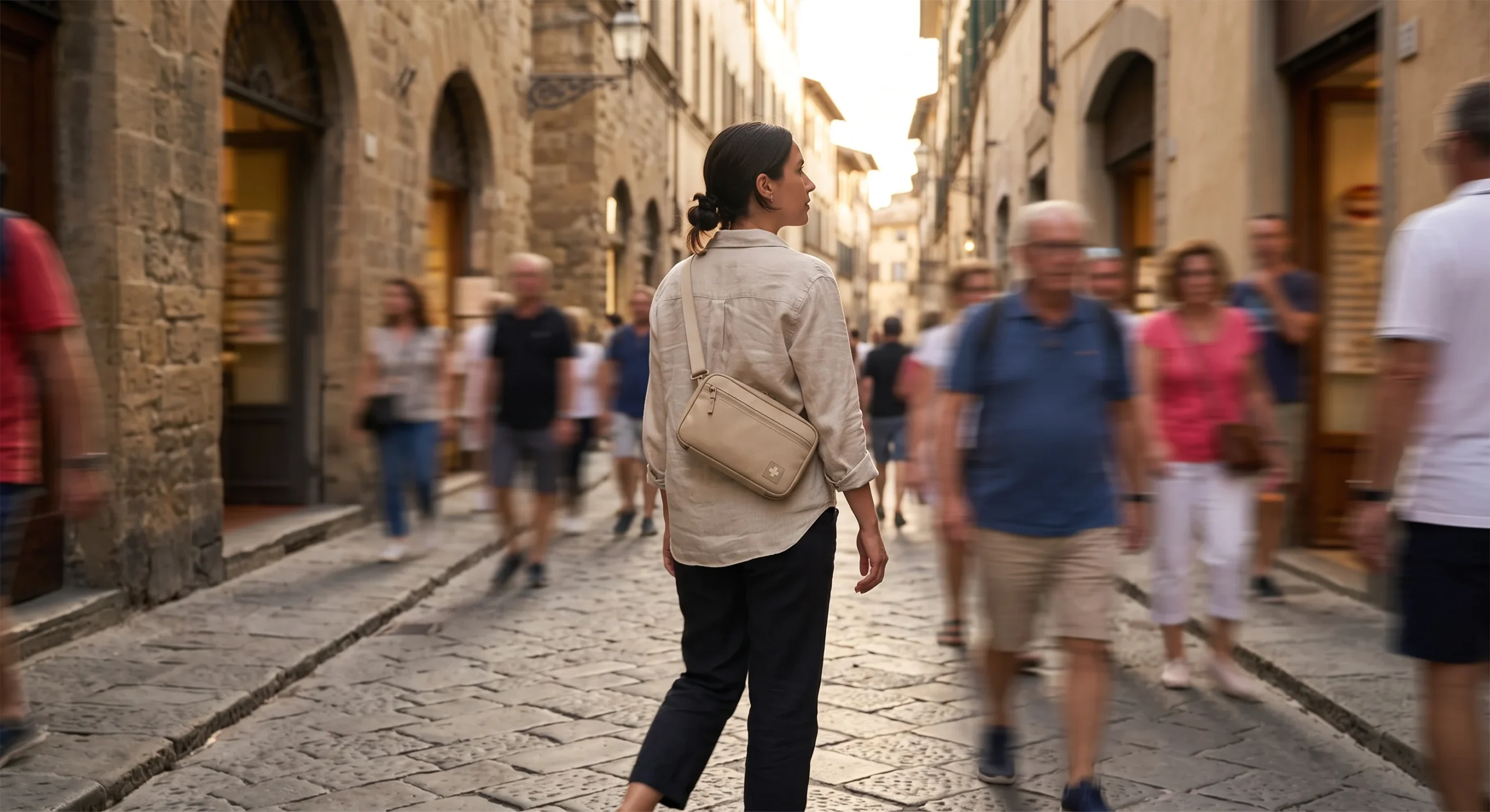 A slim crossbody daypack worn across the body of a traveler walking through a narrow European old-town street, warm afternoon light, motion blur in the background suggesting movement, candid documentary composition.