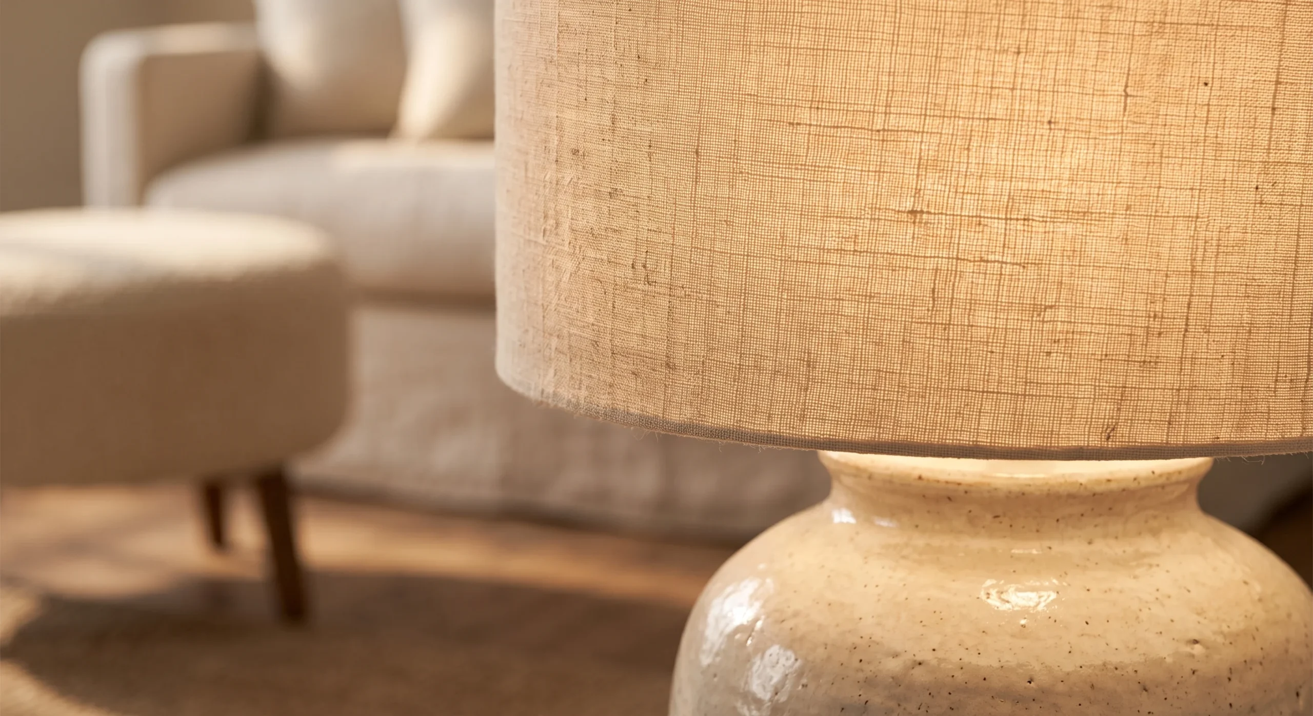 A linen drum lampshade on a ceramic lamp base, warm evening light glowing softly through the fabric, close angled shot emphasizing the linen's subtle texture illuminated from within.