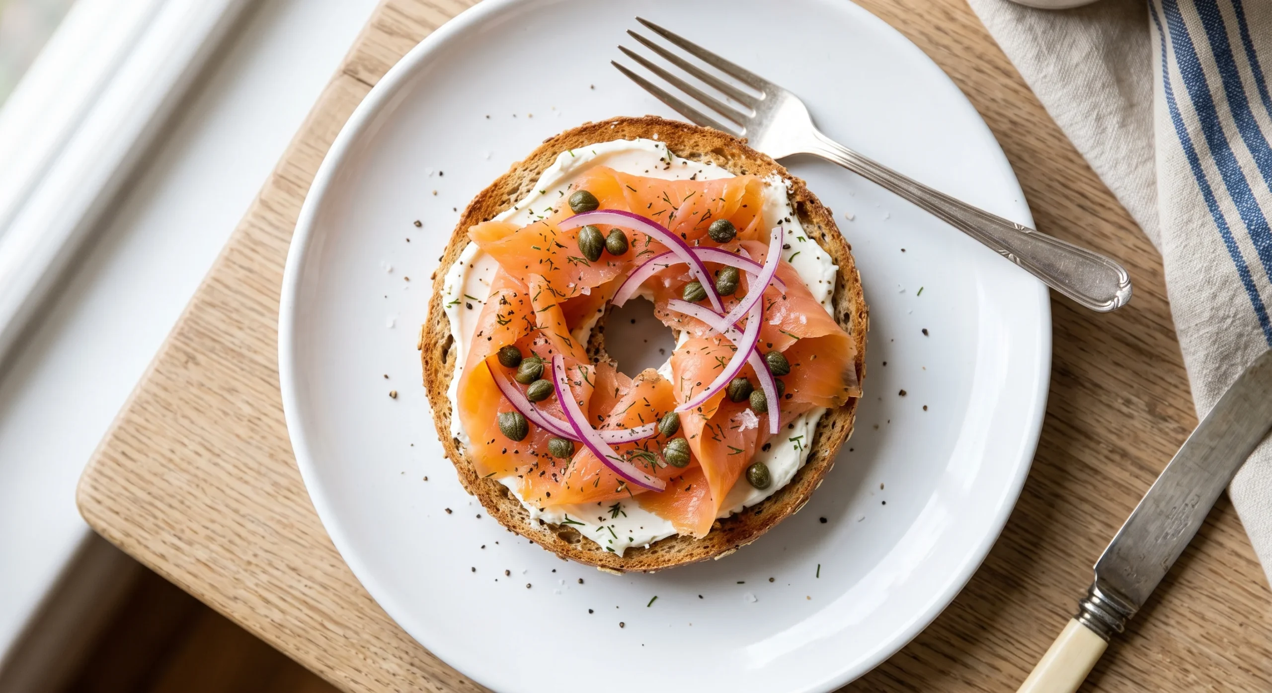 A thin bagel topped with cream cheese, smoked salmon, capers, and thin red onion slices, soft natural light, close elegant overhead composition on a simple white plate.