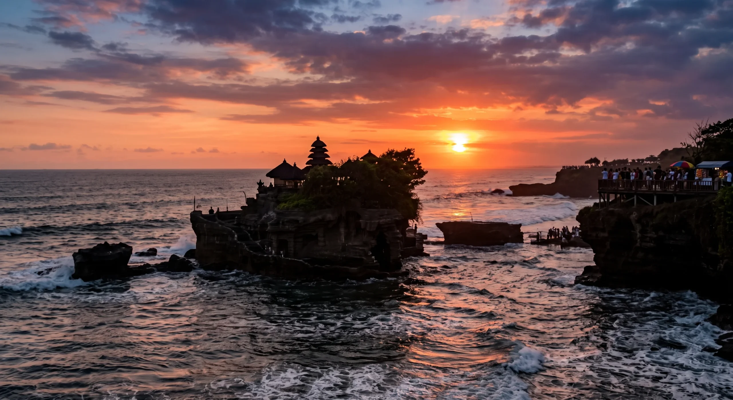Tanah Lot temple perched on an offshore rock formation fully surrounded by ocean water at high tide, silhouetted against a warm sunset sky, distant crowd gathered along a coastal viewpoint, wide dramatic seascape composition.