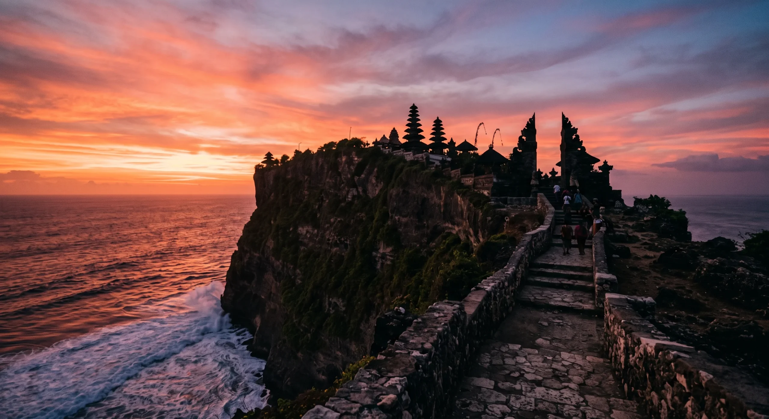 Uluwatu Temple silhouetted on a limestone clifftop against a vivid orange and pink sunset sky, ocean waves crashing far below, a narrow stone pathway leading toward the temple gate, dramatic low-angle sunset photography.