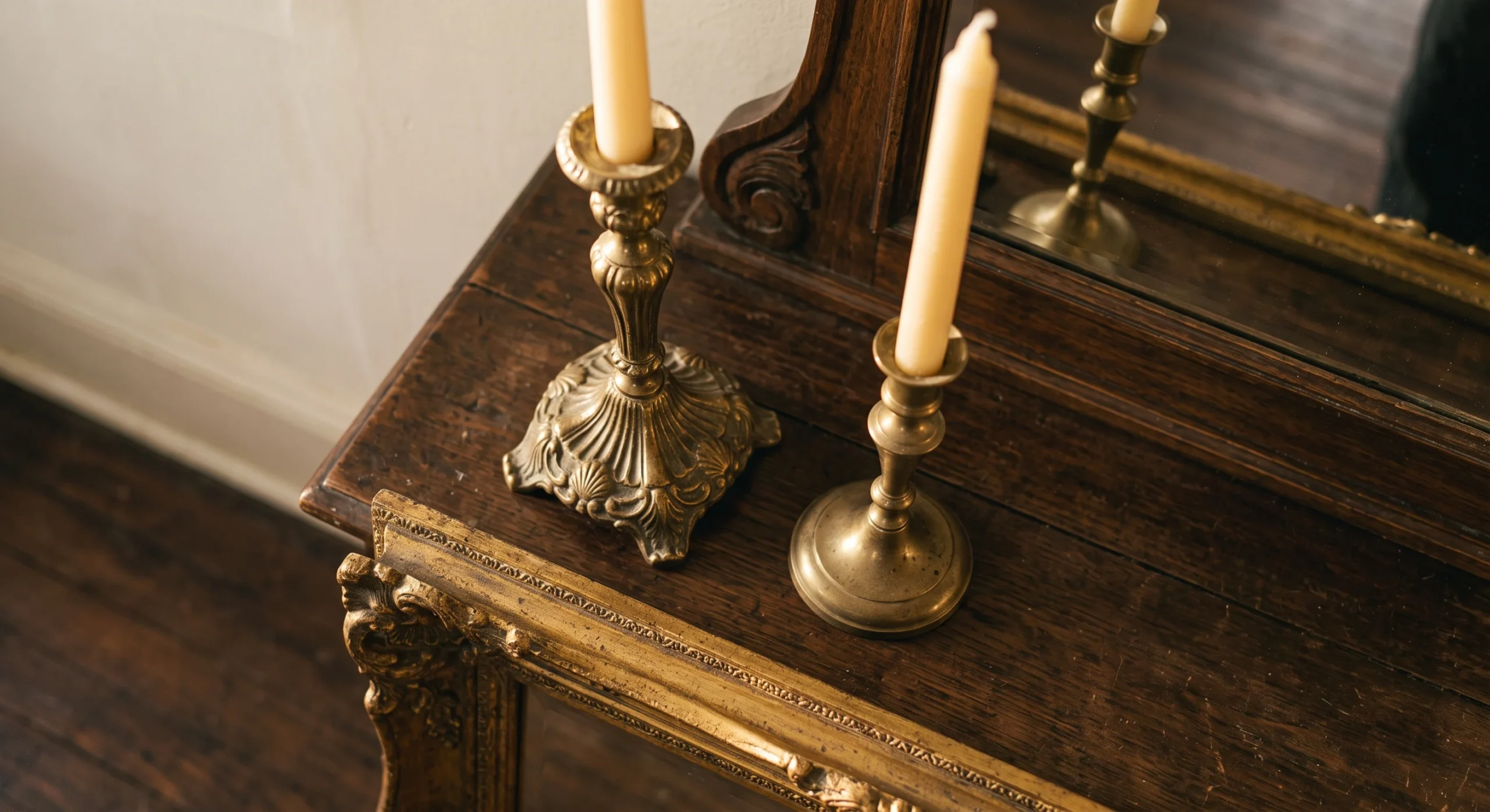 A pair of mismatched vintage brass candlesticks holding tall taper candles on a wooden console, warm evening light glowing softly across the tarnished brass surface, close overhead shot emphasizing their varied heights.